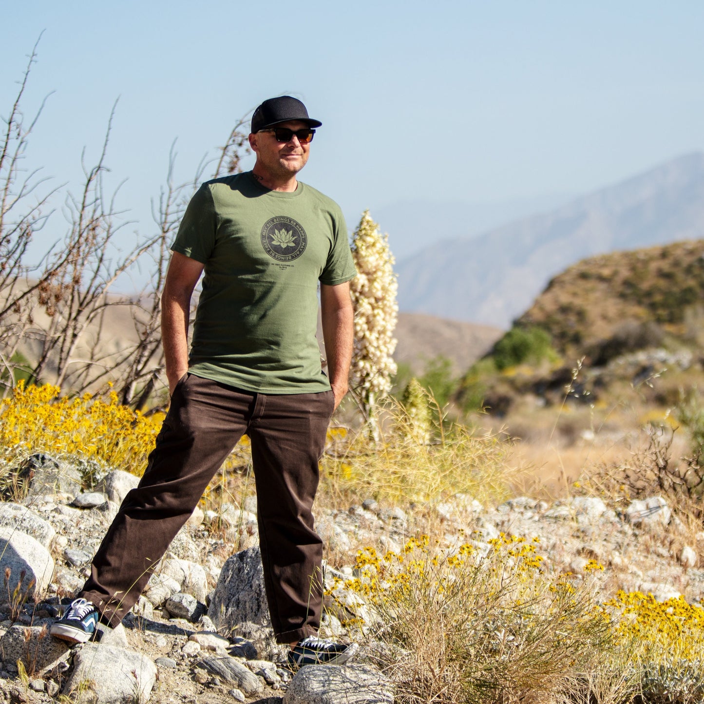 Person in a green Buddhist lotus shirt with the text, May All Beings Be Happy May All Beings Be Free, they are wearing sunglasses standing in a desert landscape with mountains in the background.