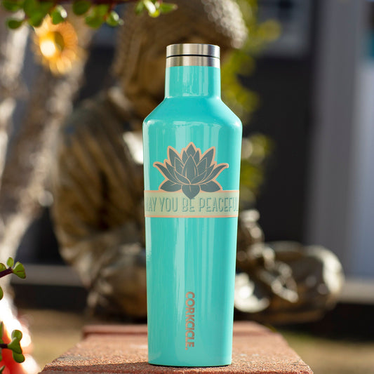 Turquoise water bottle with a lotus flower sticker with 'May you be peaceful' text on a blurred natural background.