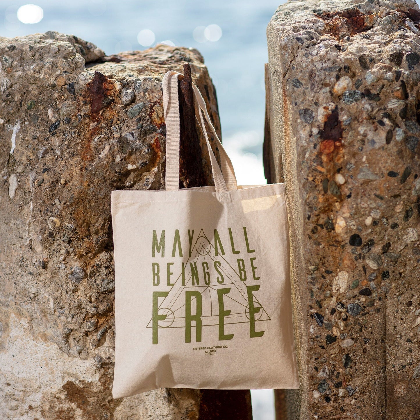 Beige tote bag with 'May All Beings Be Free' text against a textured stone wall.