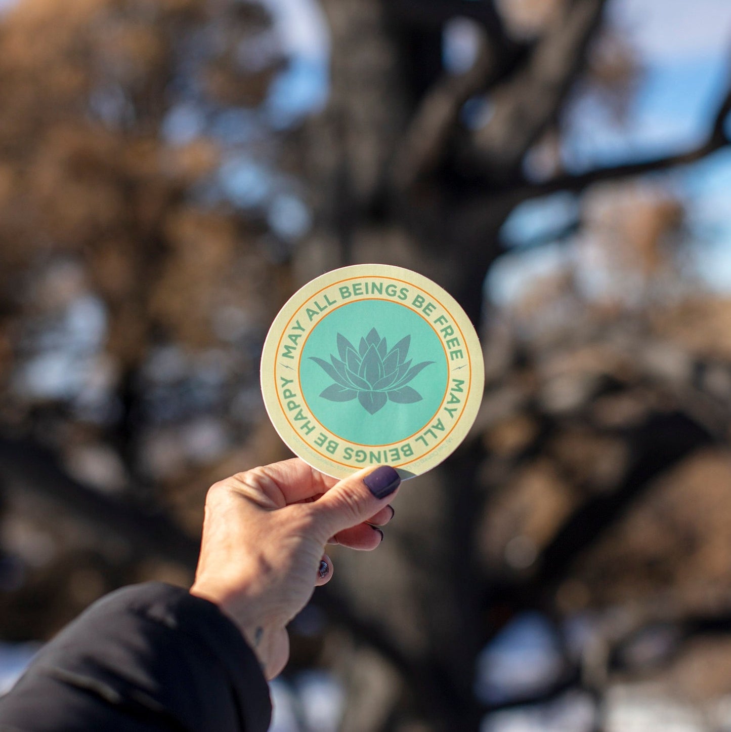 Hand holding a circular sticker with text may all beings be happy may all beings be free and a lotus flower design against a blurred natural background.