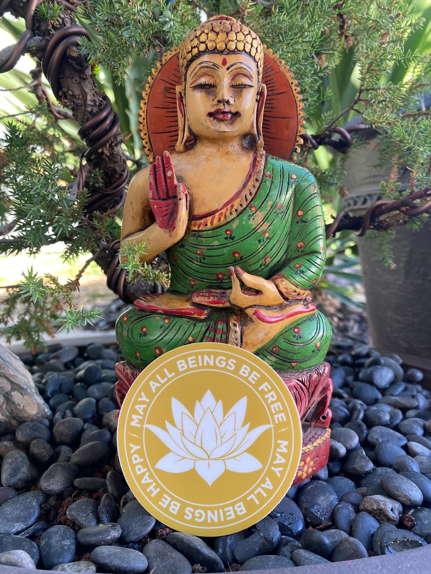 Colorful Buddha statue with a 3" gold and white sticker with a lotus flower design and text, may all beings be free may all beings be happy, on pebbles and plants.