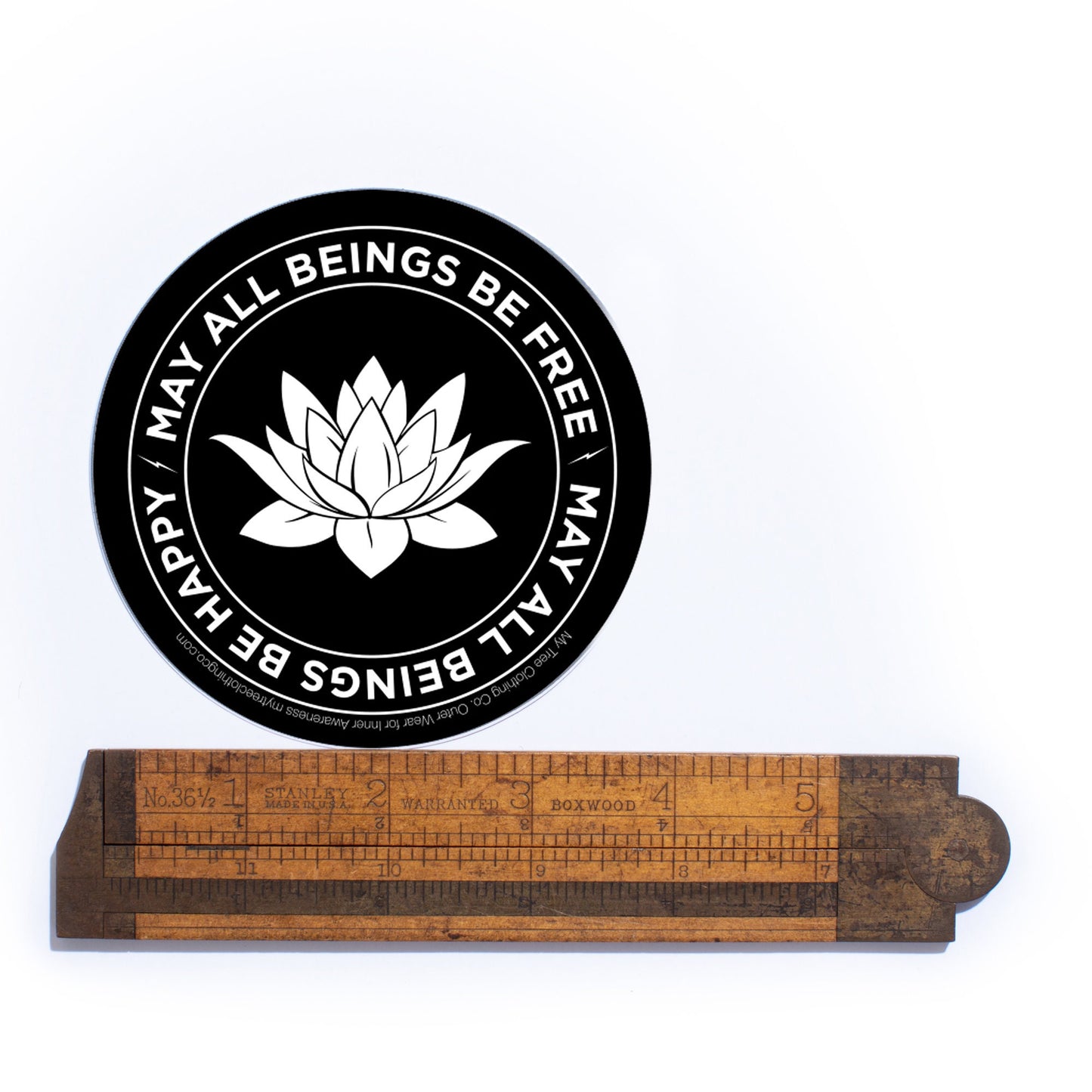 4" black and white circular sticker with a lotus flower design and text, MAY ALL BEINGS BE FREE MAY ALL BEINGS BE HAPPY placed above a wooden ruler on a white background.