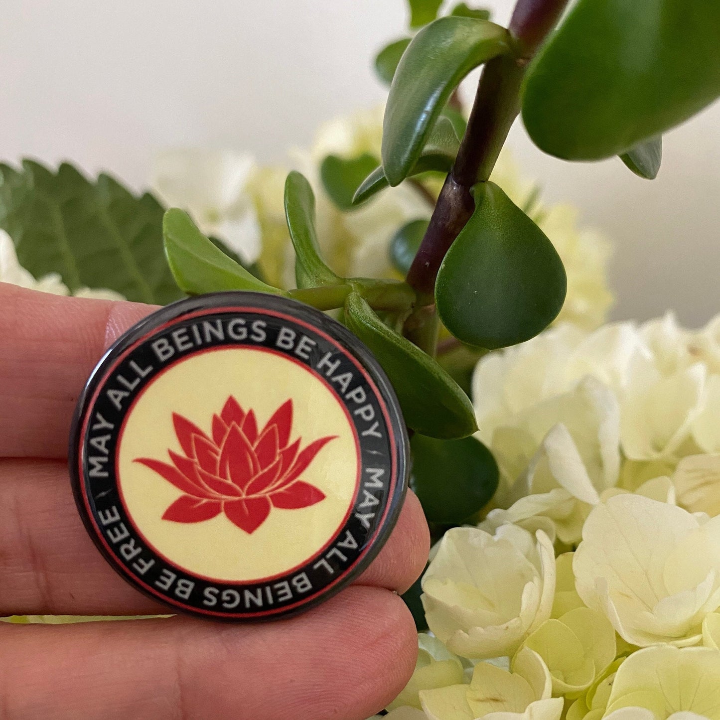 1.25" pin back button with a red lotus flower design and the text, may all beings be free may all beings be happy, held by a hand against a floral background.
