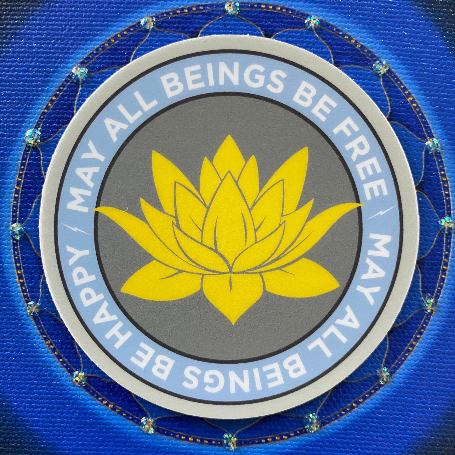 3" circular sticker with a yellow lotus flower and text 'MAY ALL BEINGS BE FREE, HAPPY, HARMONY' on a blue background.