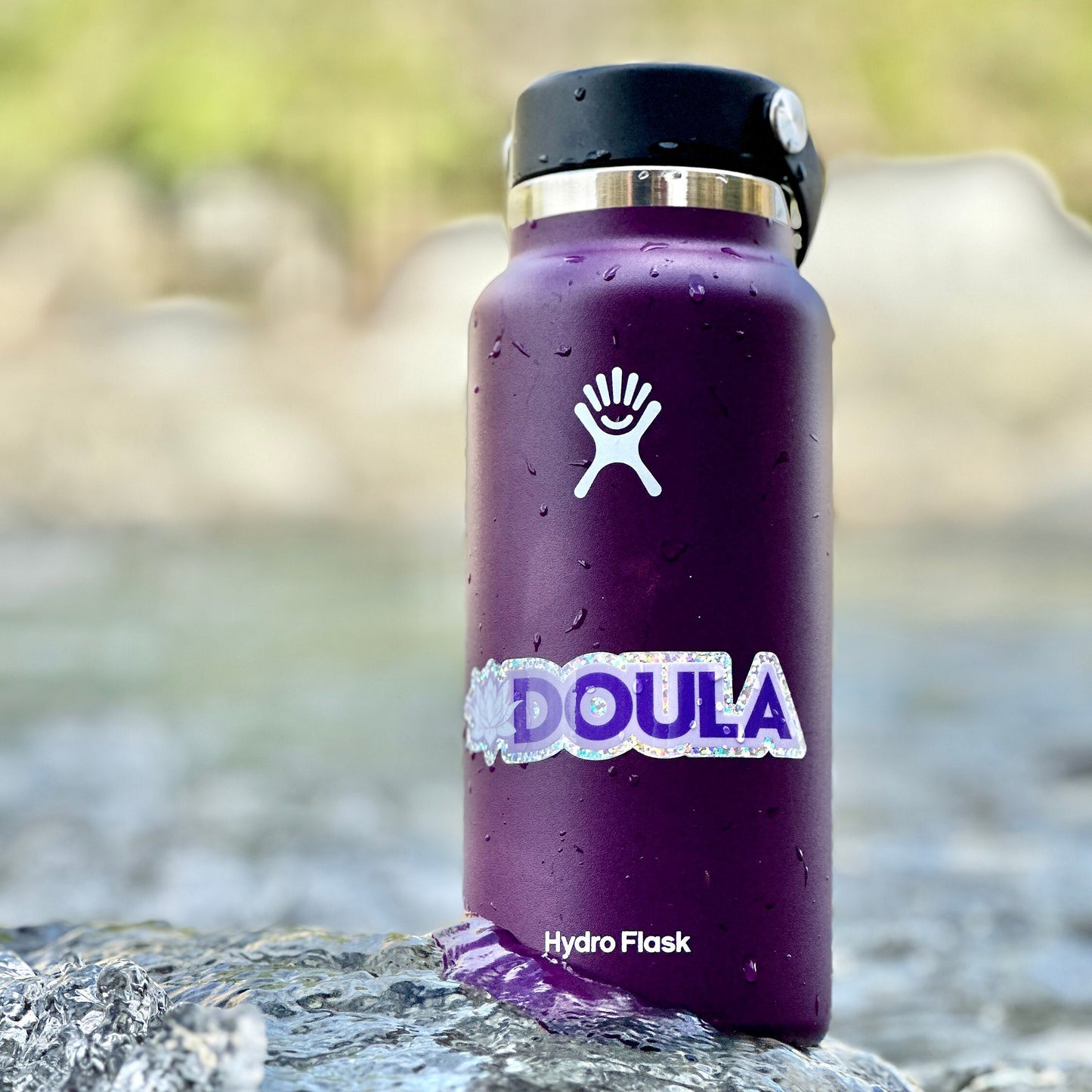 Purple Hydro Flask water bottle with 'DOULA' sticker on a rock.