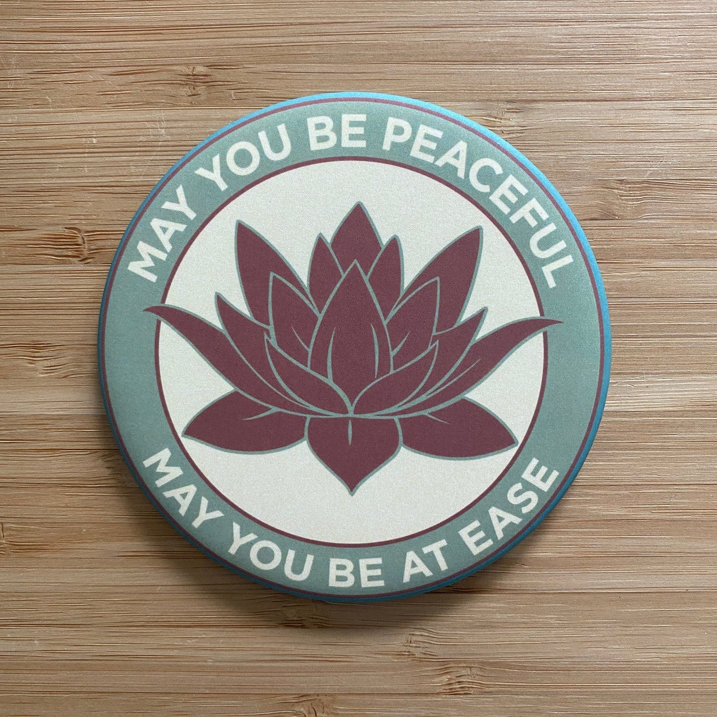 Round Buddhist mantra magnet with lotus flower design and text 'May you be peaceful, may you be at ease' on a wooden surface.