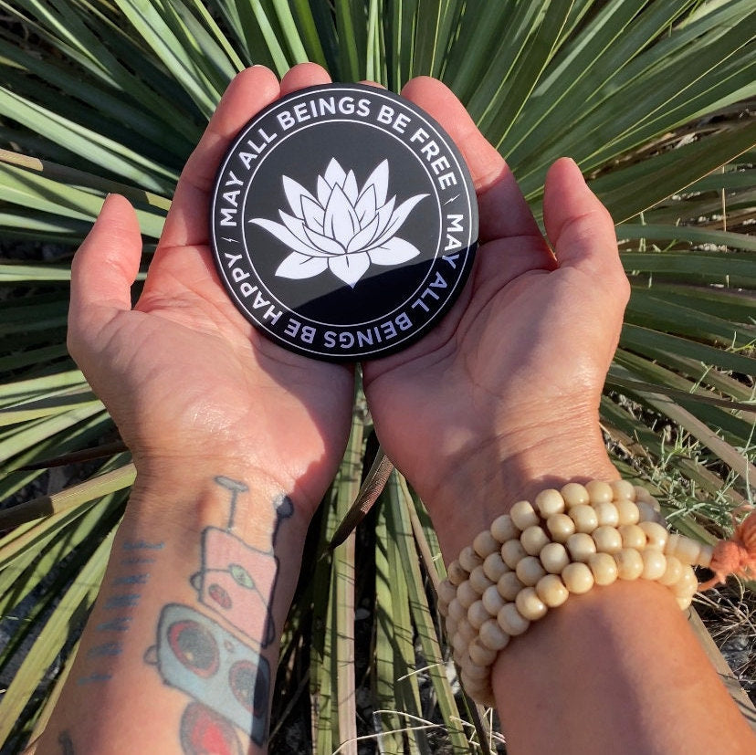 Person holding a 3.5" round black and white magnet with lotus flower design and text, may all beings be free, may all beings be happy, against a natural background.
