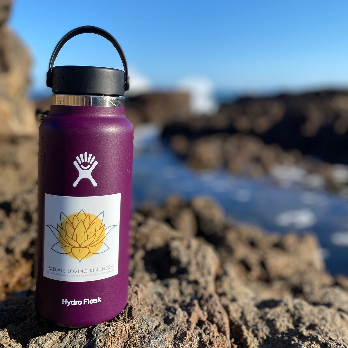 Purple Hydro Flask water bottle with a yellow flower 'Radiate Loving Kindness' 3" square sticker on a rocky surface.