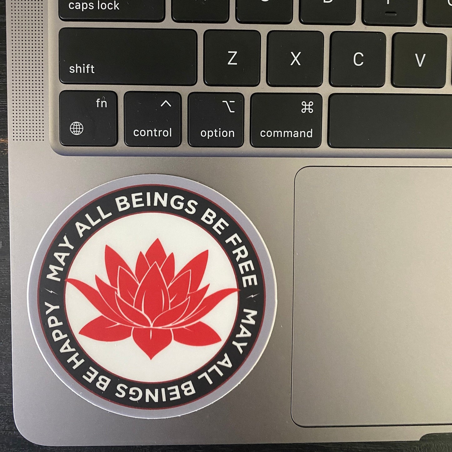 Laptop with a red and white sticker featuring a lotus flower and text, may all beings be free may all beings be happy, on the palm rest area.