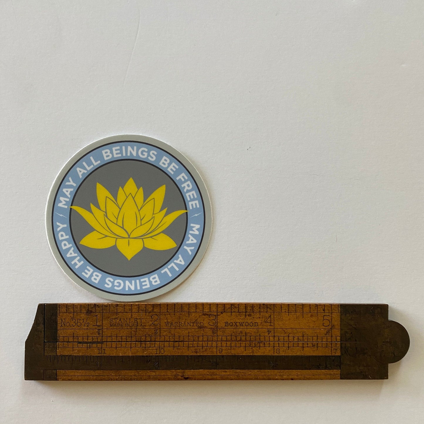 3" circular sticker with a lotus flower design and text, may all beings be free may all beings be happy, next to a wooden ruler for scale.