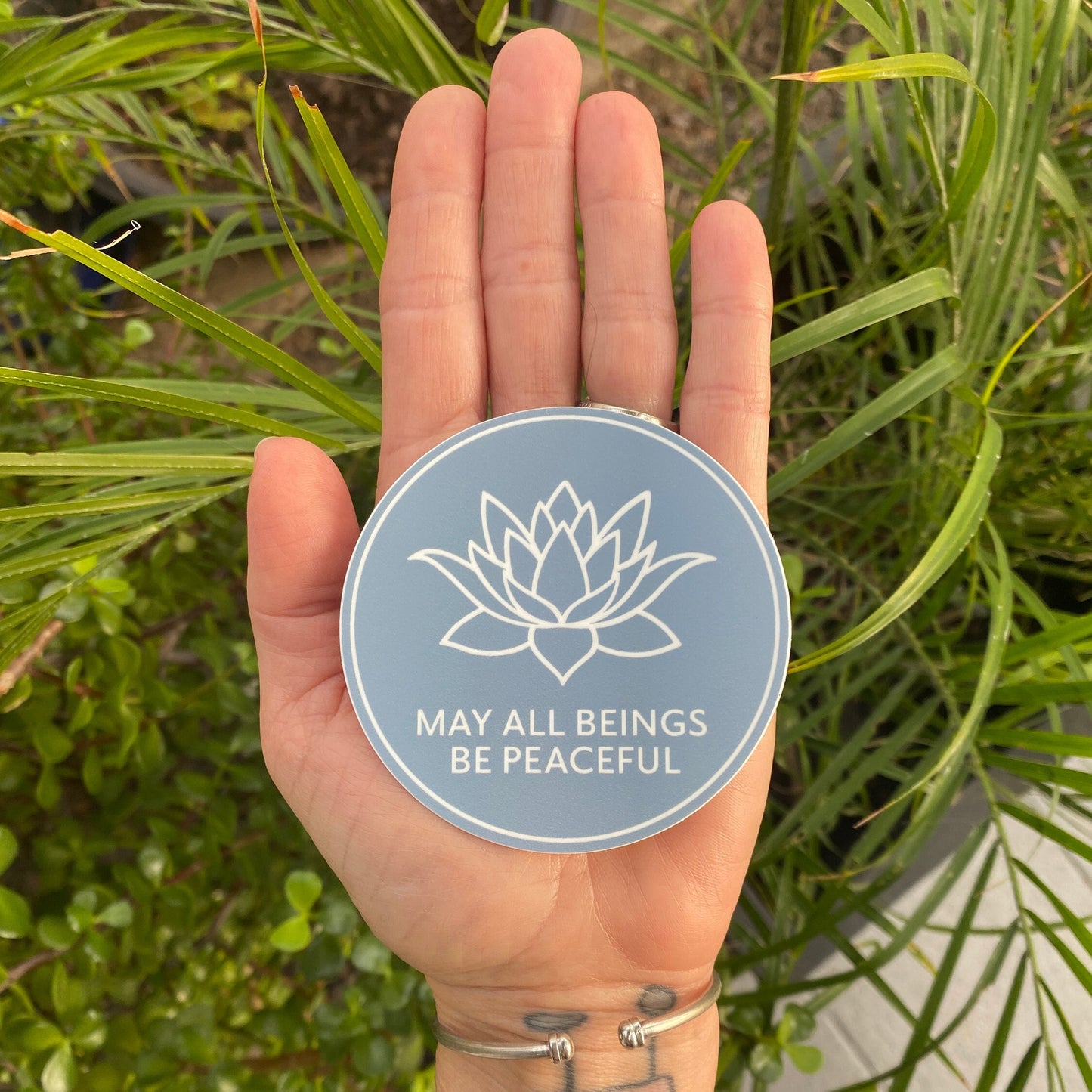 Hand holding a round 3" blue sticker with a lotus flower design and text, MAY ALL BEINGS BE PEACEFUL, against a green plant background