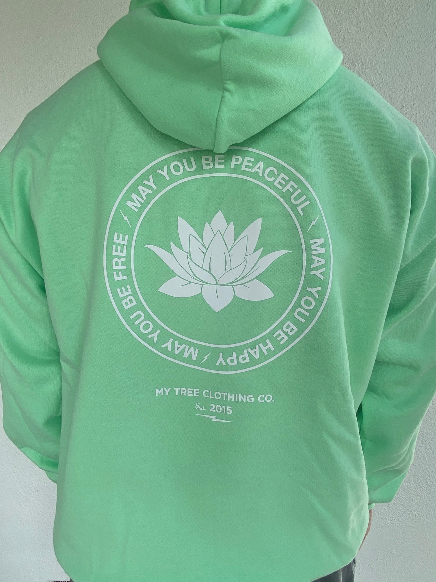 Green hoodie with a white lotus flower design and text may you be peaceful may you be happy may you be free on the back against a plain background.