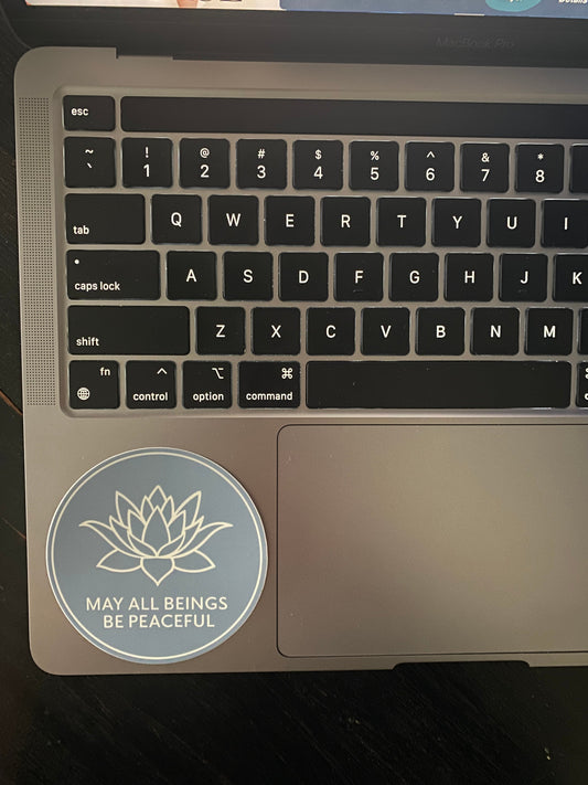 Laptop keyboard with a sticker featuring a round 3" blue sticker with a lotus flower design and text, MAY ALL BEINGS BE PEACEFUL.
