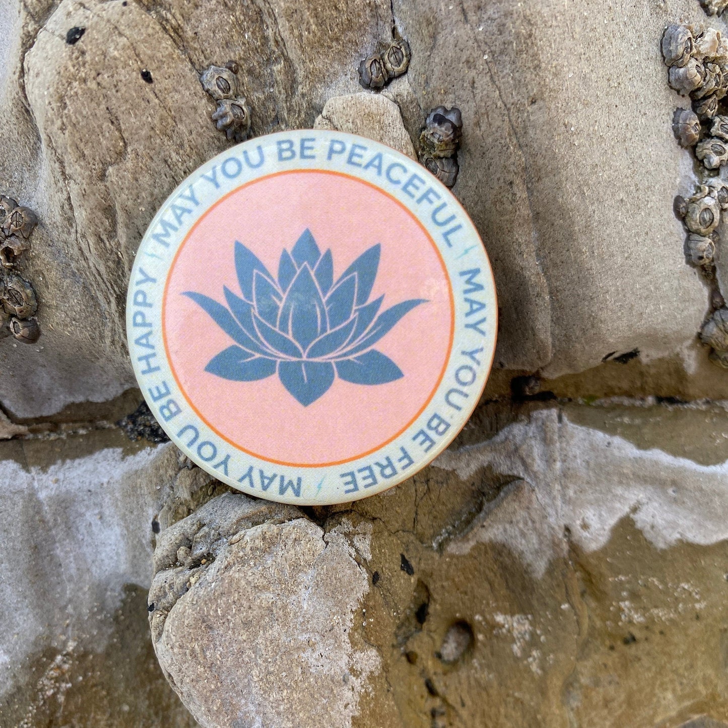 Round pink and blue sticker with lotus flower design and the text, may you be peaceful may you be happy may you be free, on textured stone surface