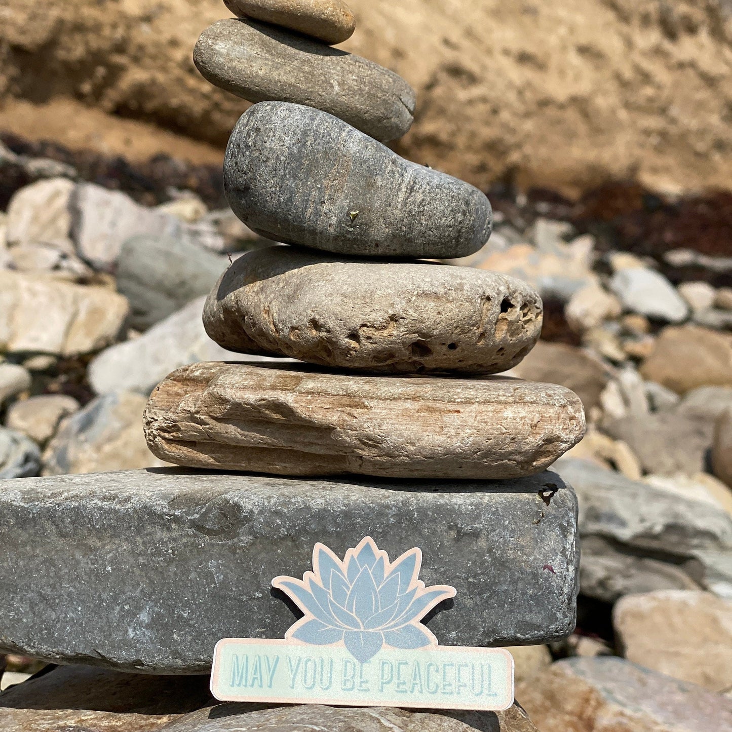 Stack of stones with a 'May You Be Peaceful' sticker on a rocky background.