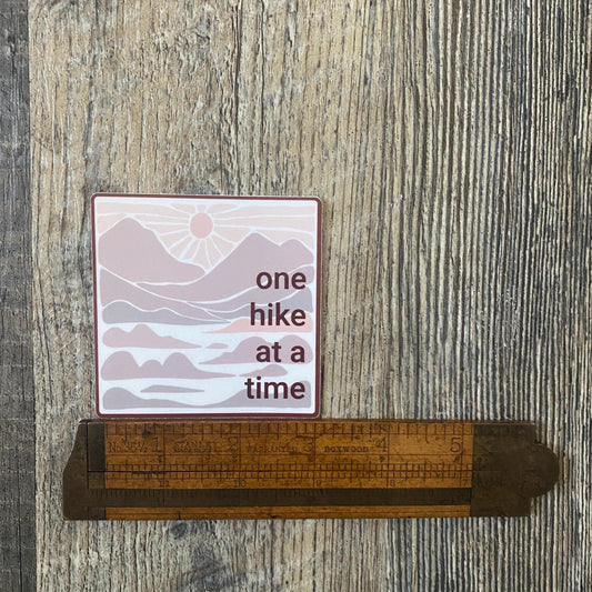 3" square sticker with 'one hike at a time' in neutral colors with mountains and a sunset on a wooden surface with a ruler for scale.
