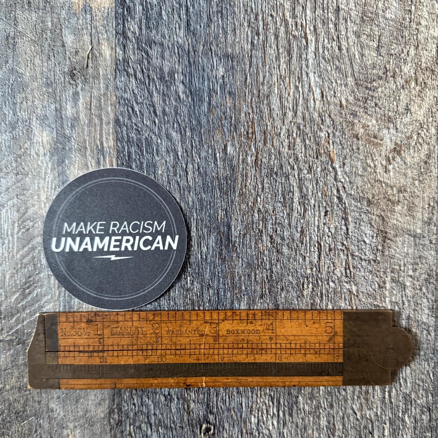 Circular sticker with 'Make Racism Unamerican' text on a wooden ruler against a textured gray background being measured by a wooden ruler.