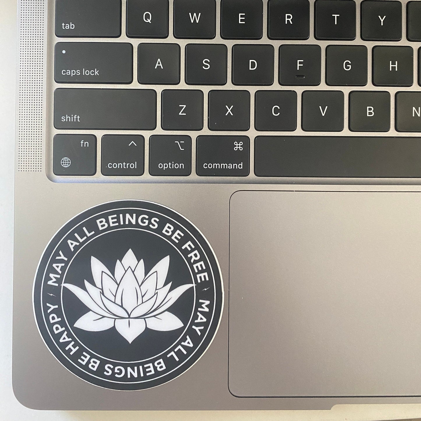 Laptop keyboard with a 3" circular sticker with a white lotus flower design and text, may all beings be free may all beings be happy, on the palm rest area.