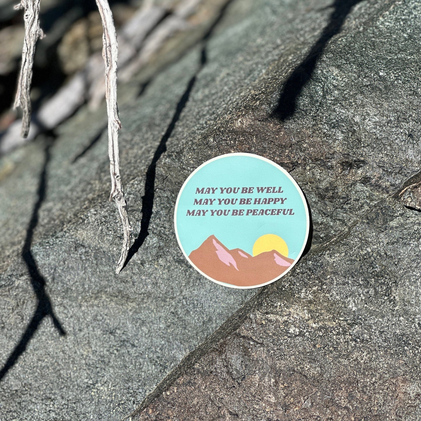 Circular Buddhist 3" sticker design featuring mountains and text, may you be well may you be happy may you be peaceful, on a rock in the mountains of southern California.