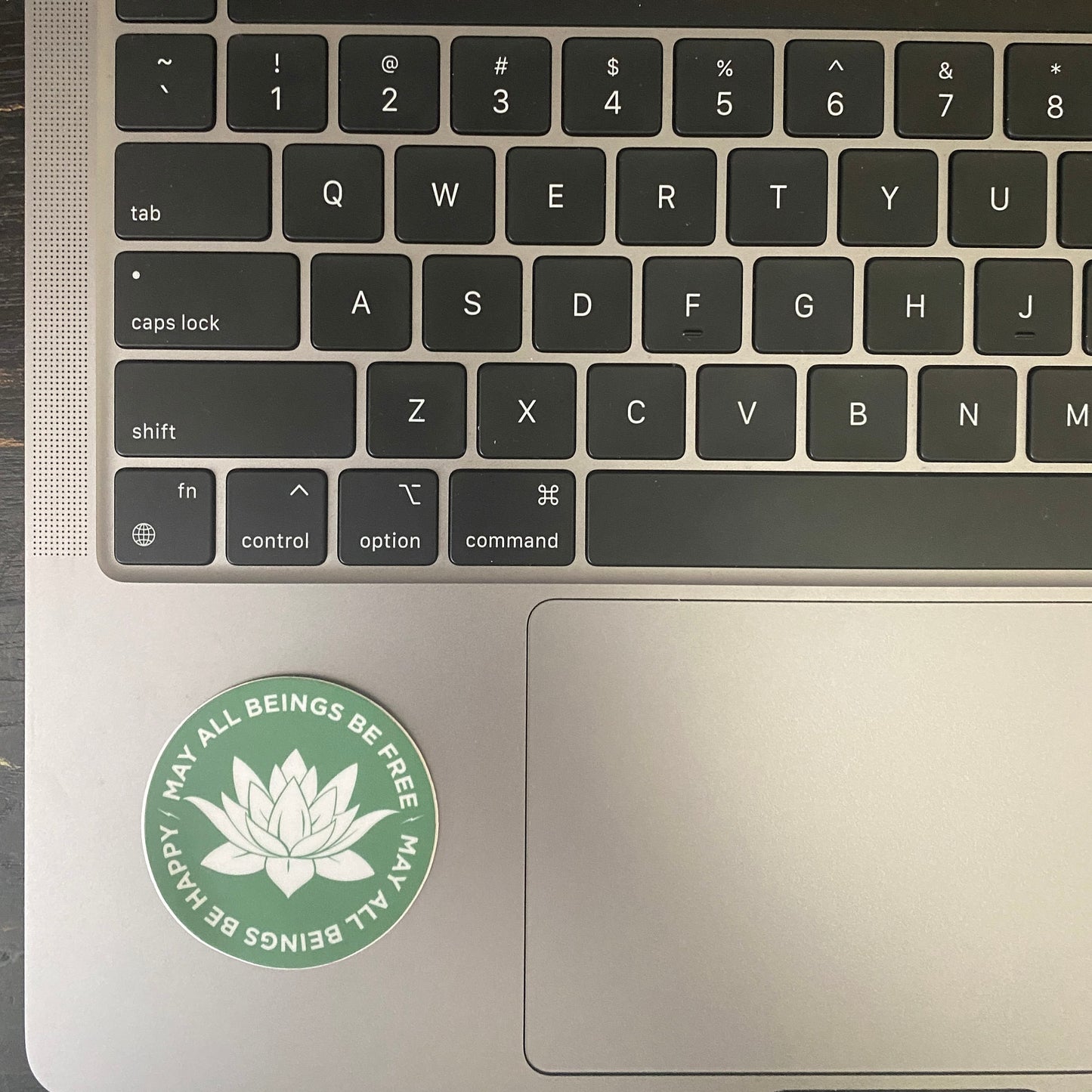 Laptop keyboard with a 2" round green sticker with a white lotus Buddhist design and the text, may all beings be free may all beings be happy.