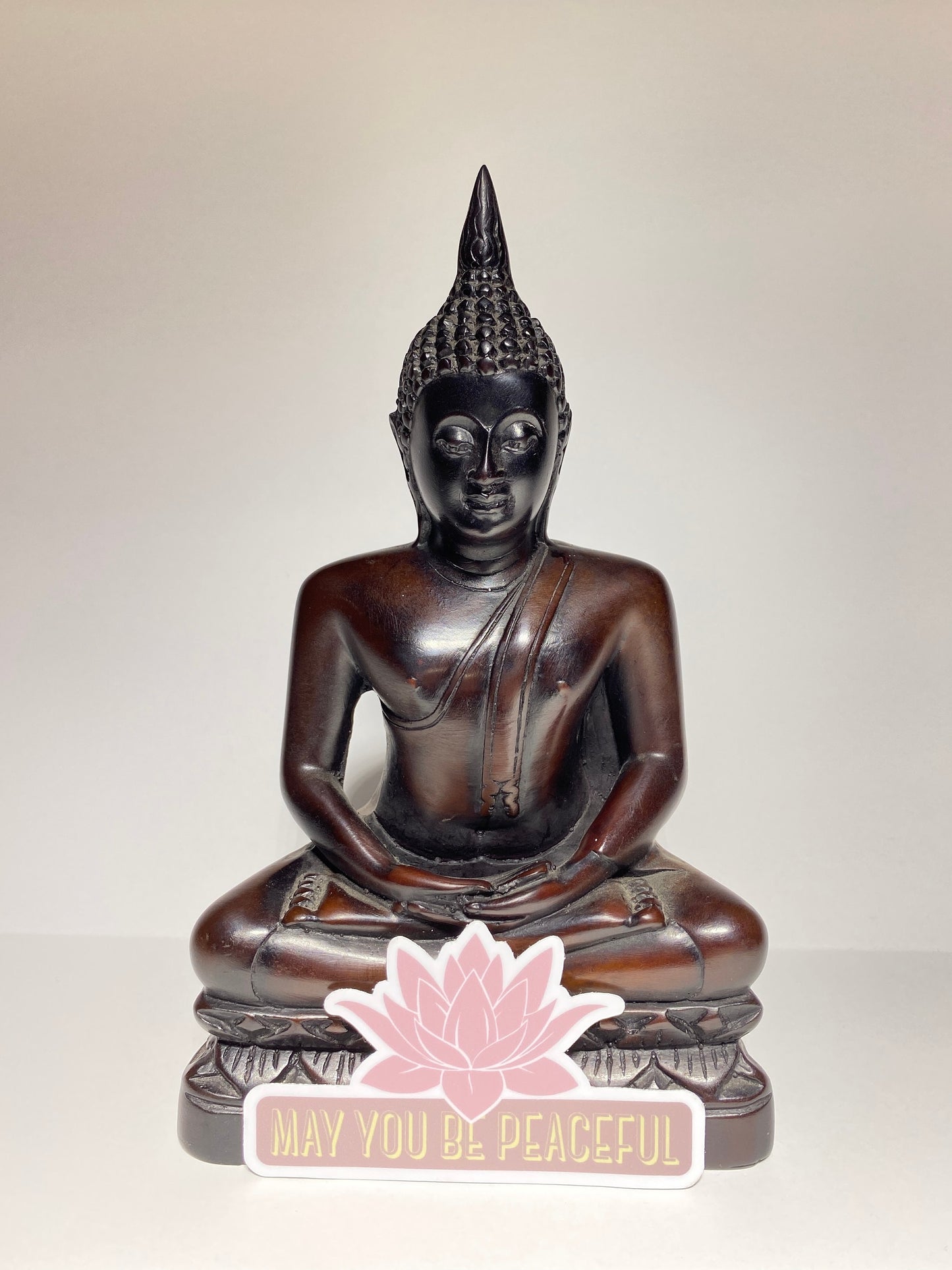 Wooden Buddha statue with a pink lotus and 'May you be peaceful' text on a beige background.