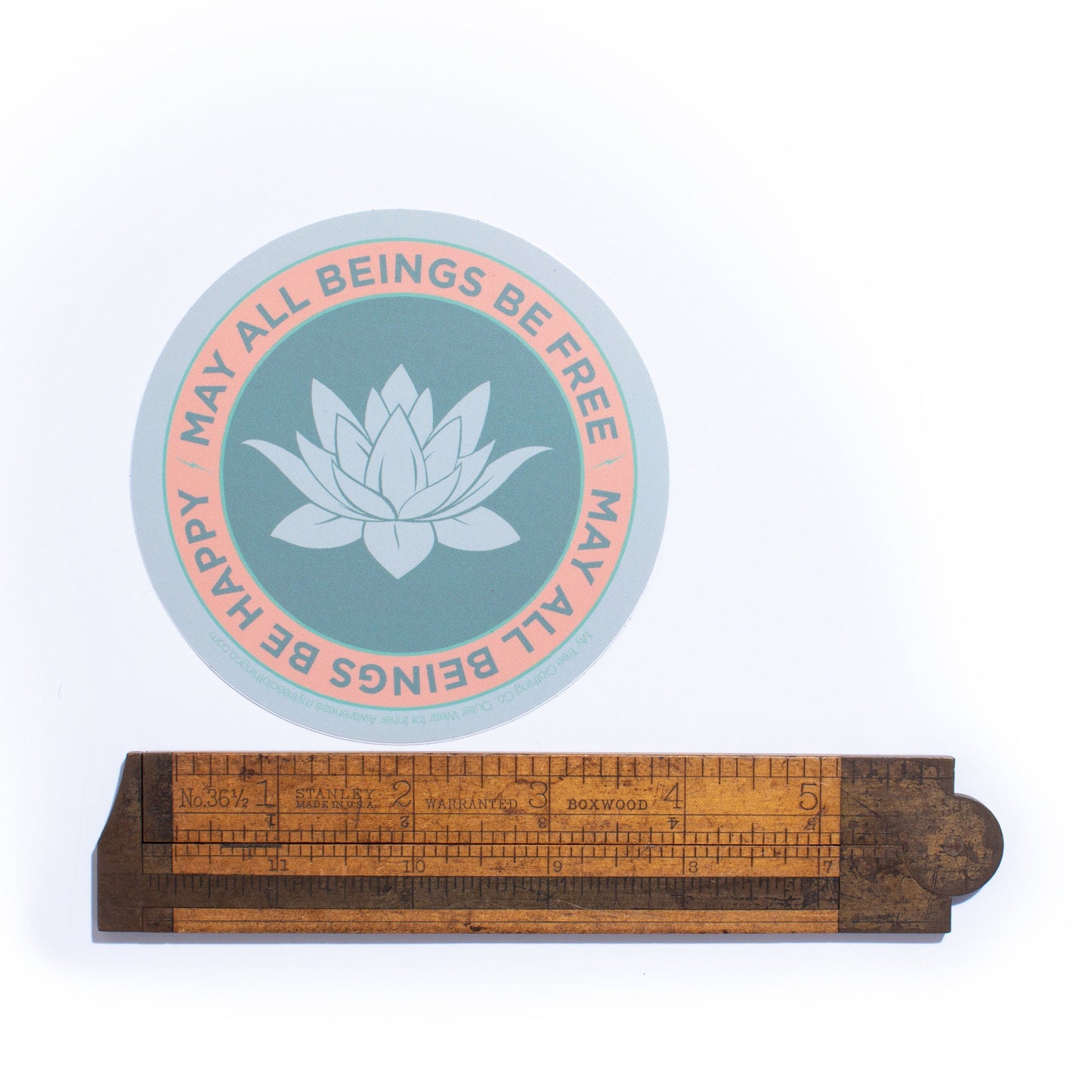 4" circular sticker with a lotus flower design and text, may all beings be free may all beings be happy, on a white background, accompanied by a wooden ruler.