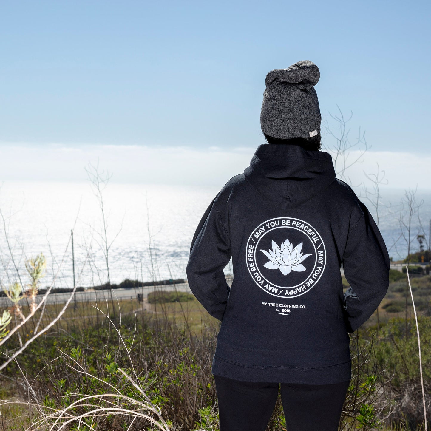 Backside view of a person wearing a black hoodie with a white lotus and the mettā prayer, 'may you be peaceful may you be happy may you be free.'standing outdoors with a scenic background.