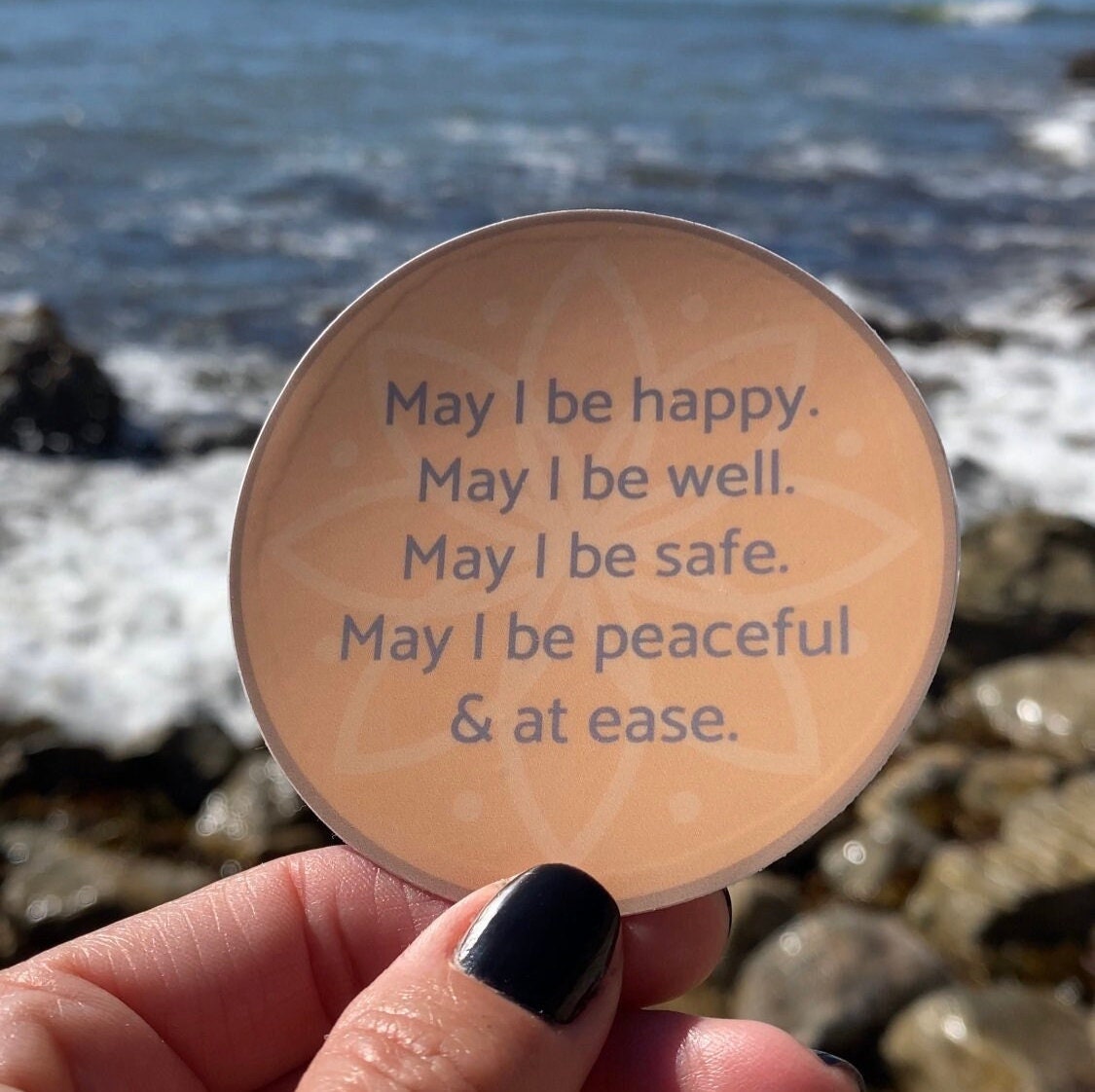 Three inch round circular sticker with text, may i be happy may i be well may i be safe may i be peaceful may i be at ease, held in a hand in front of the Pacific Ocean.