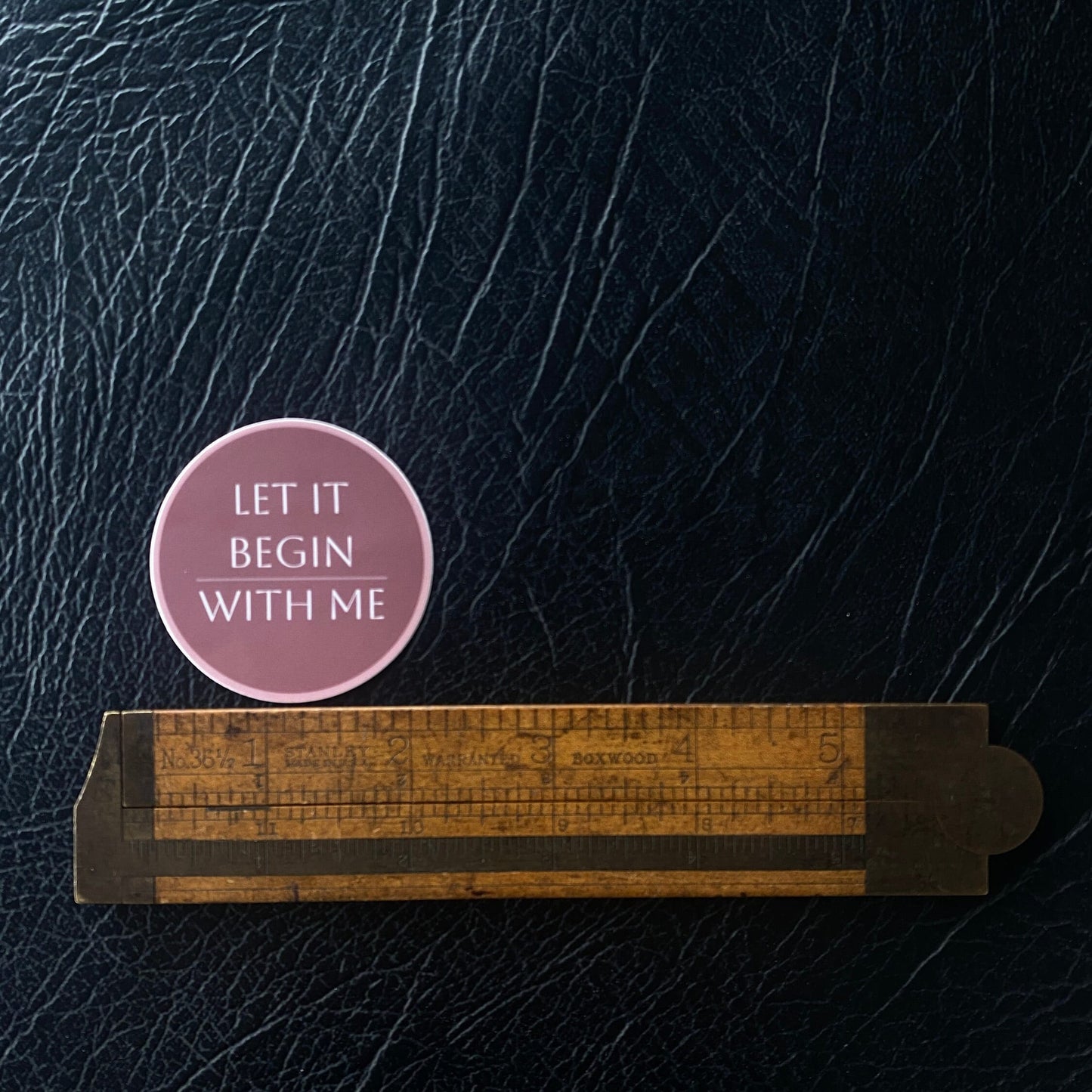 Pink 2" circular sticker with 'LET IT BEGIN WITH ME' text on a textured dark surface.