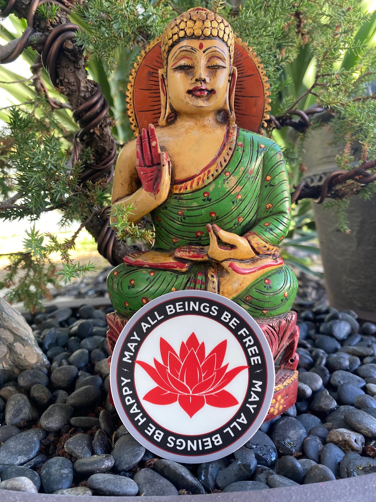 Statue of a seated Buddha figure with a red and white 3" sticker featuring a lotus flower and metta prayer, may all beings be free may all beings be happy, on a pebble surface.
