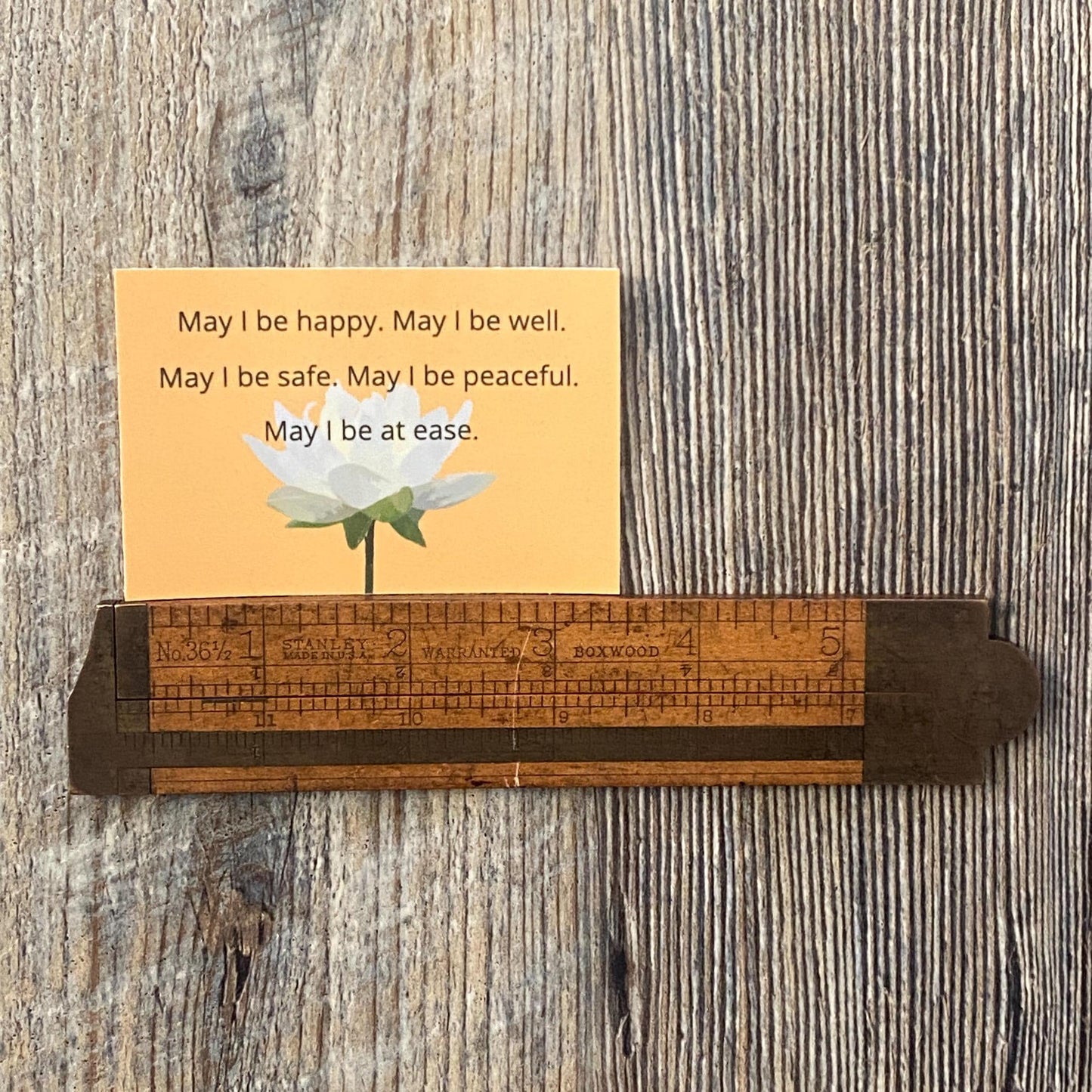 Wooden ruler with a rectangular yellow sticker with a lotus and the words, may i be happy may i be well may i be safe may i be peaceful on a wooden surface.