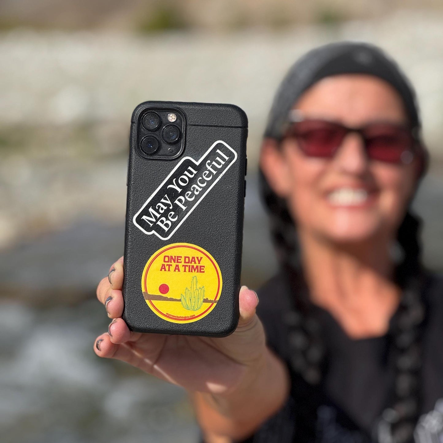 Person holding a phone case with two inch round sticker that reads, one day at a time, outdoors.
