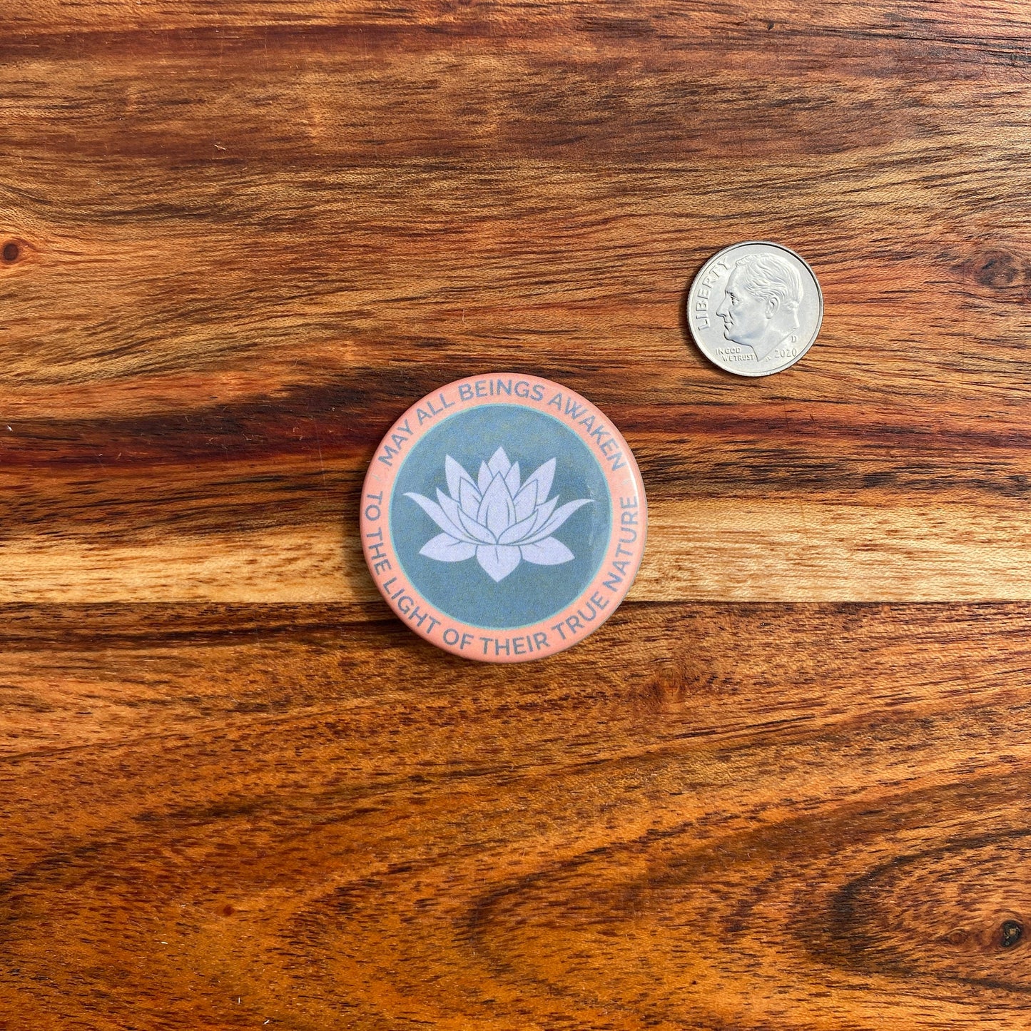 Circular button with a lotus flower design and text, may all beings awaken to the light of their true nature, on a wooden surface next to a coin for scale.