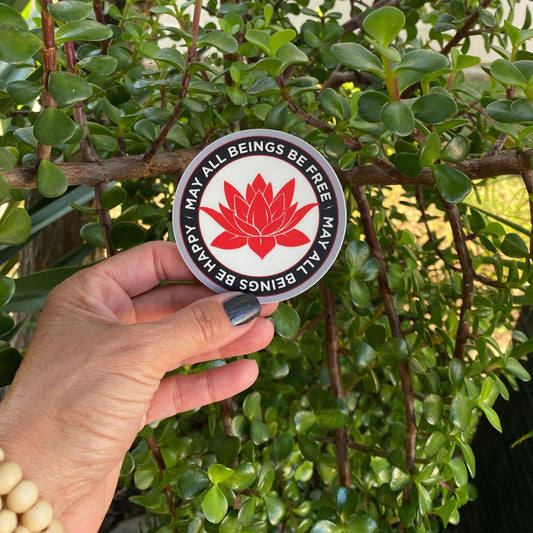 Person holding a circular sticker with a red lotus flower design and text, may all beings be free may all beings be happy, against a green plant.