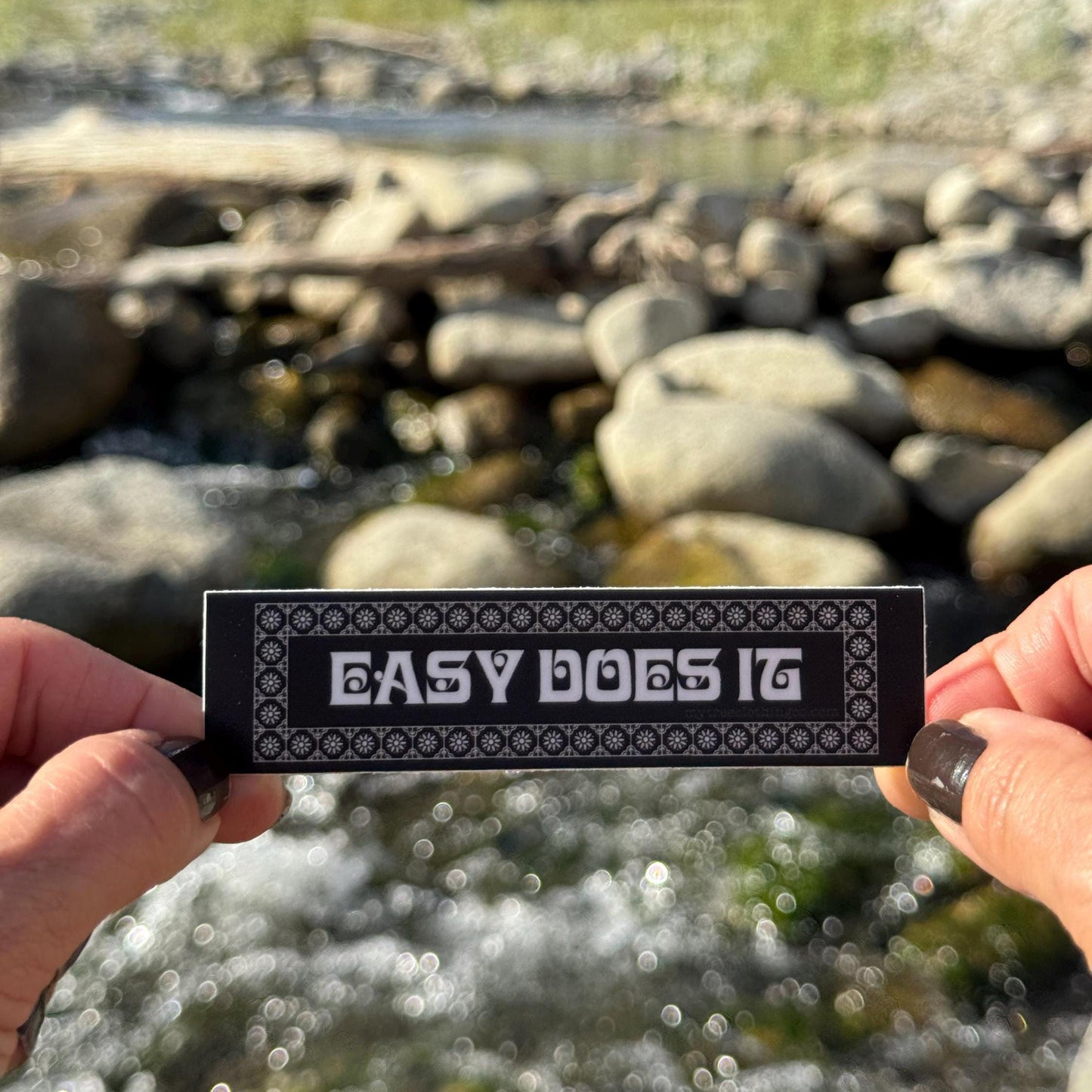 Person holding a 'Easy Does It' sticker with a natural background of rocks and water.