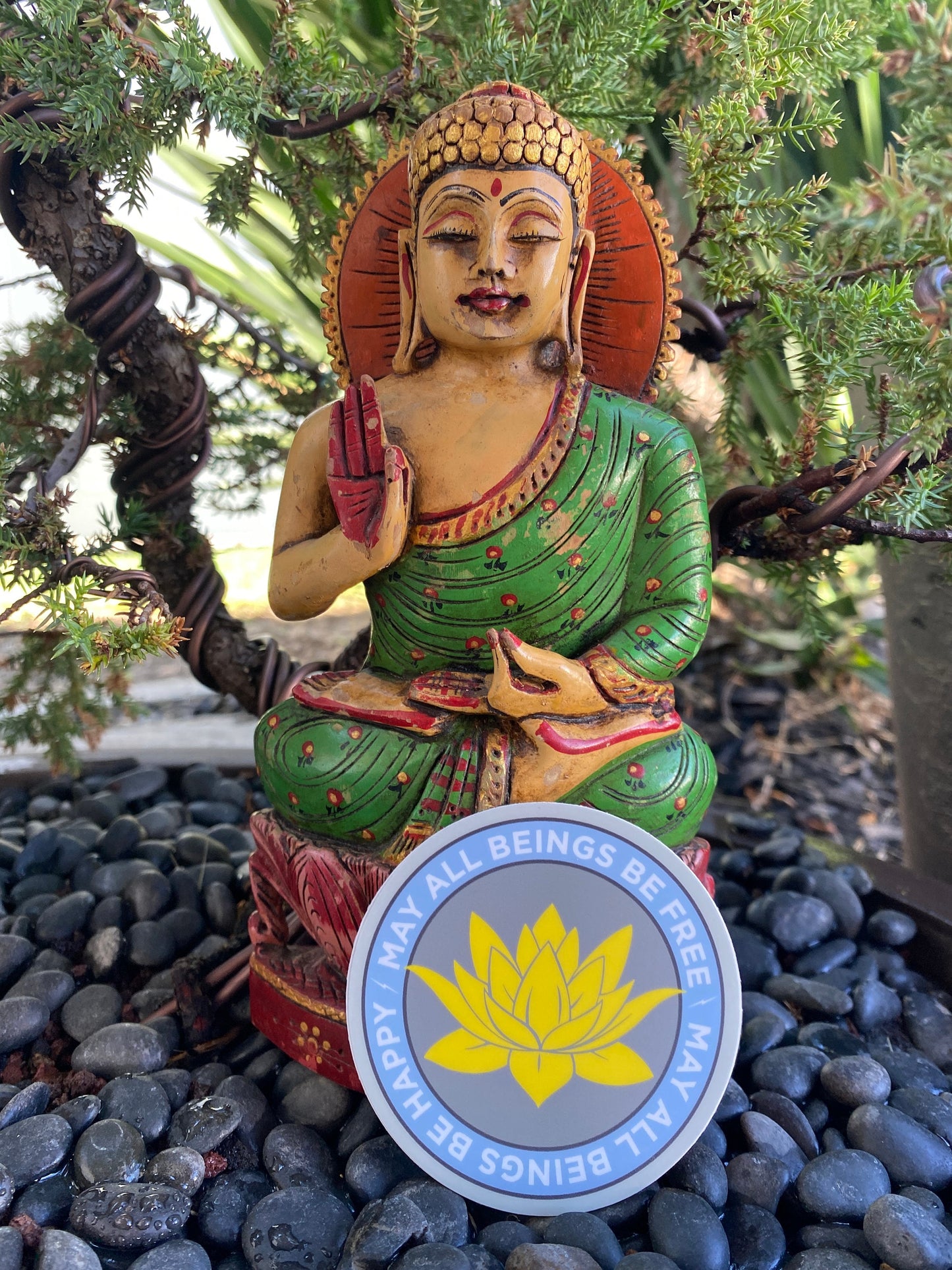 Statue of a seated Buddha figure with a 3" circular sticker with a lotus flower design and text, may all beings be free may all beings be happy on pebbles and greenery.