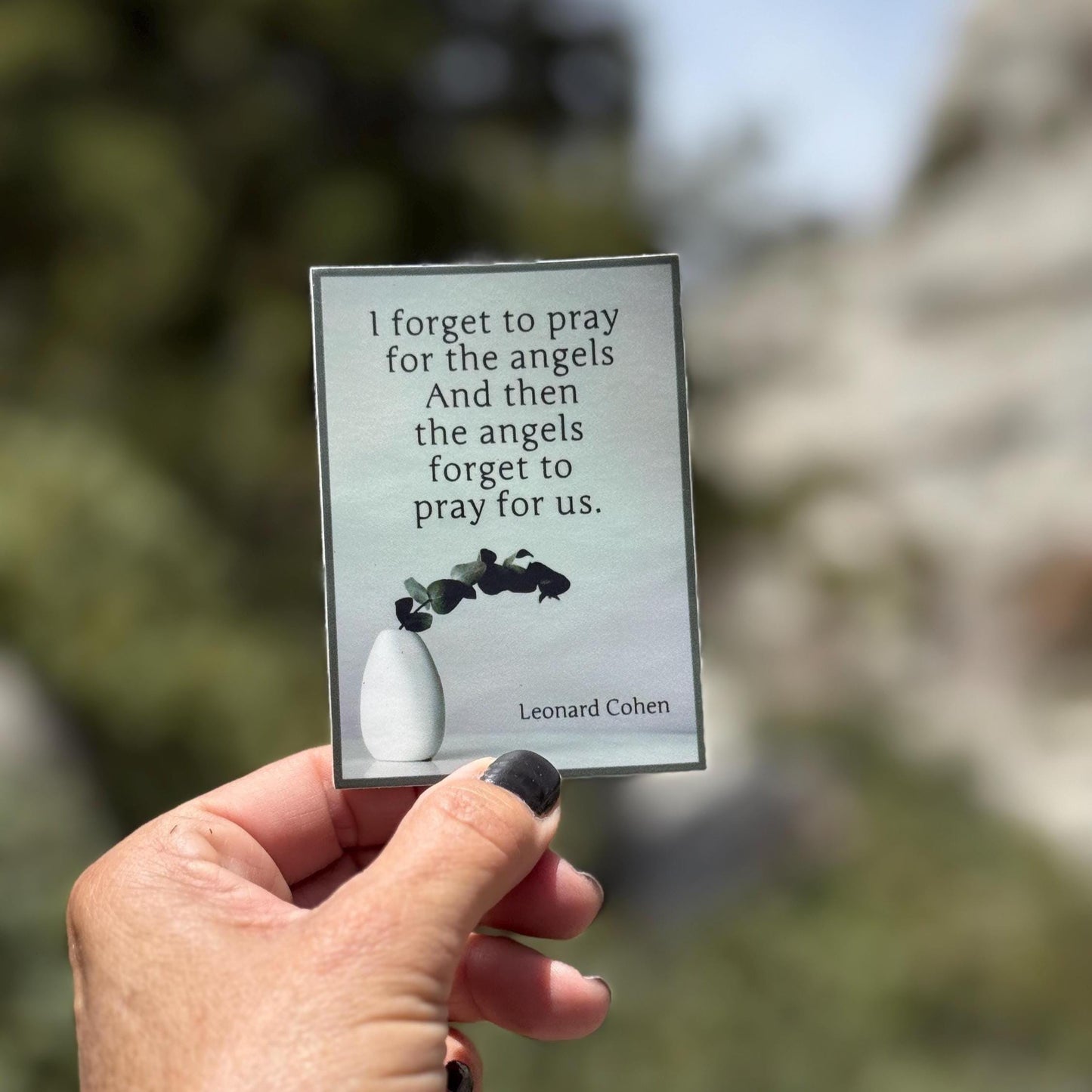 Hand holding a sticker of a Leonard Cohen quote, 'I forget to pray for the angels and then the angels forget to pray for us.'