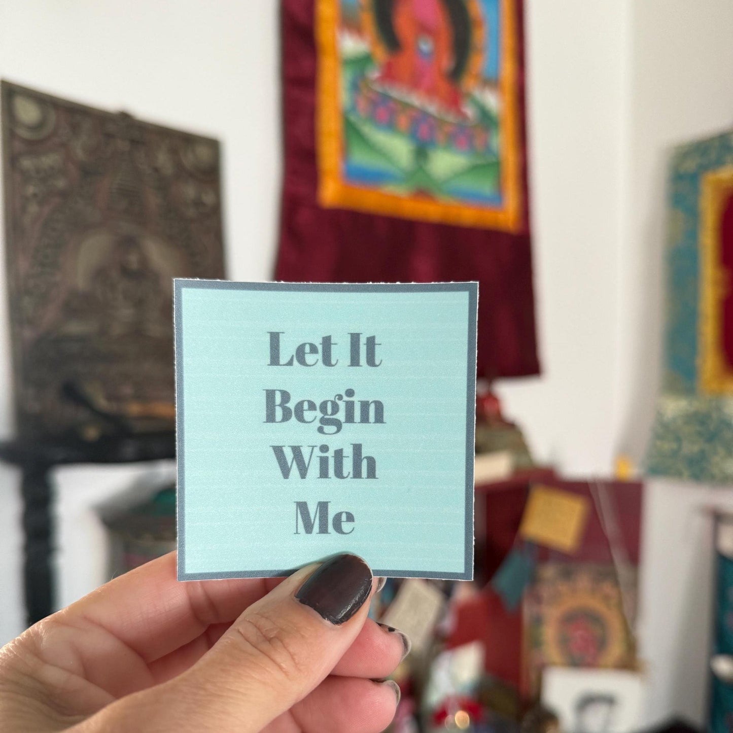 Hand holding a blue, 2" square sticker with 'Let It Begin With Me' text in front of colorful wall art.