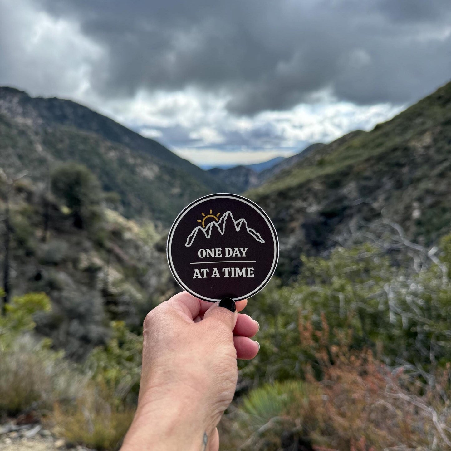 Hand holding a "3 round sticker with outline of mountains and a sunset with 'One Day at a Time' text with a mountainous landscape in the background