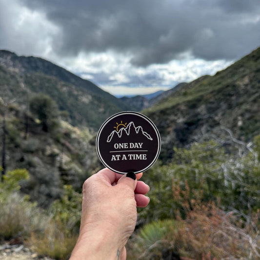 Hand holding a "3 round sticker with outline of mountains and a sunset with 'One Day at a Time' text with a mountainous landscape in the background