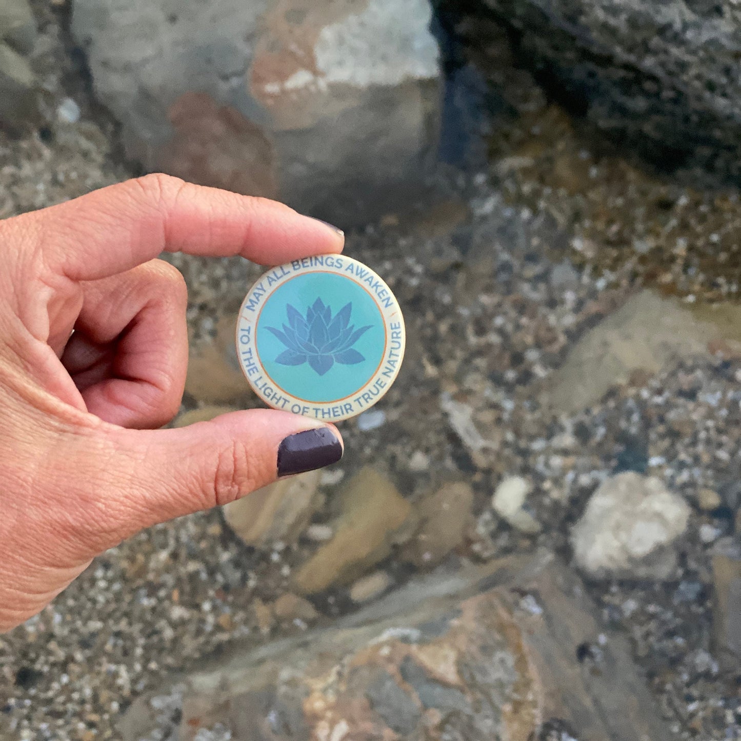 Hand holding a 1.5" button with the mantra that reads, may all beings awaken to the light of their true nature, with a lotus design against a natural background.
