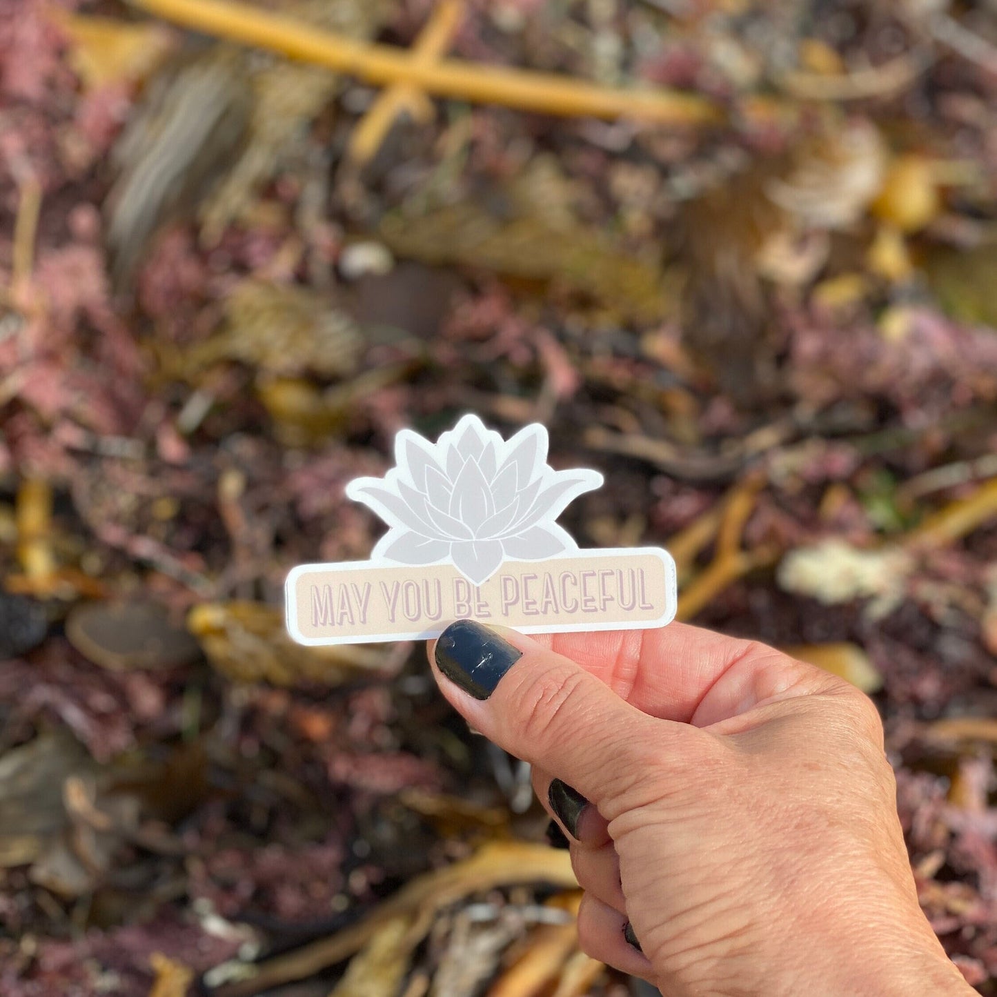 Hand holding a pale lavender lotus design die cut vinyl sticker with the text, may you be peaceful.