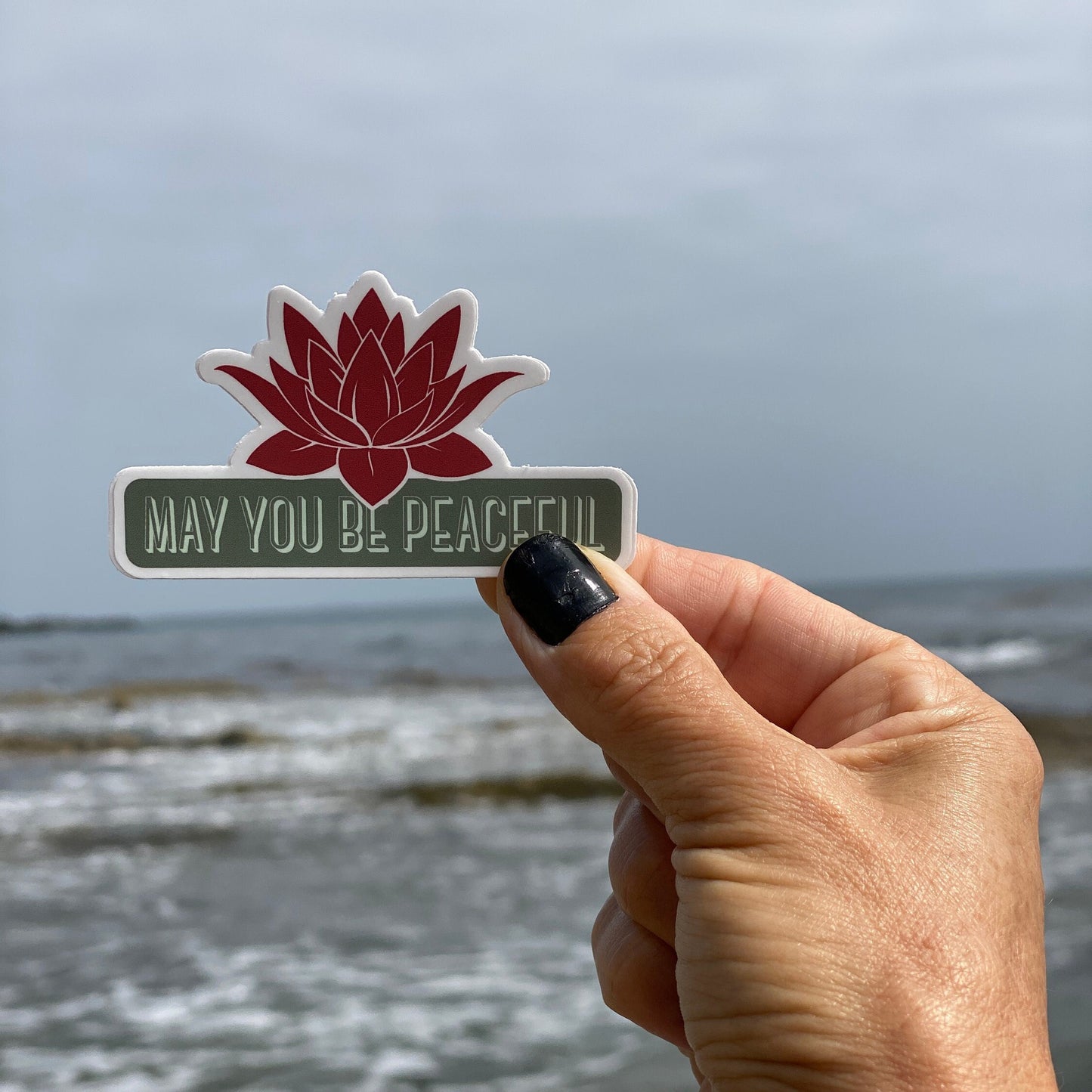 Hand holding a sticker with a lotus flower design and 'MAY YOU BE PEACEFUL' text against a beach background.