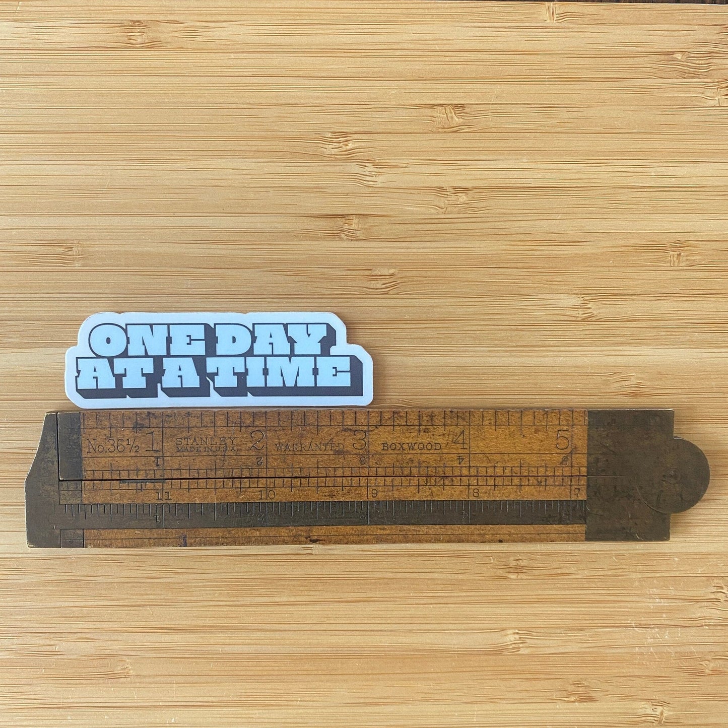 Sticker with 'ONE DAY AT A TIME' text on a wooden surface with a ruler.
