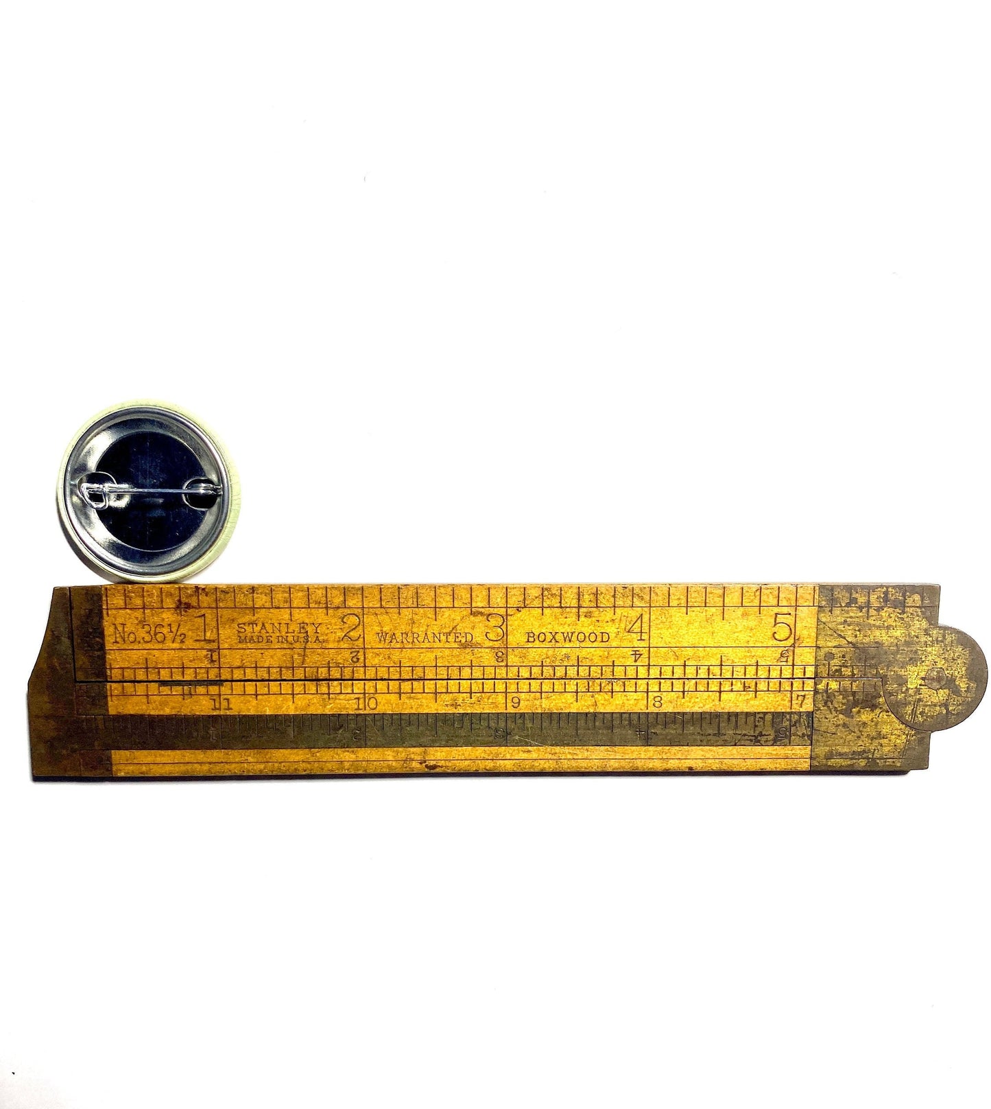 1.25" Orange round pin back button with a buddhist lotus flower and the text 'may all beings be happy may all beings be free' being measured by an antique wooden ruler with measurement markings on a white background.