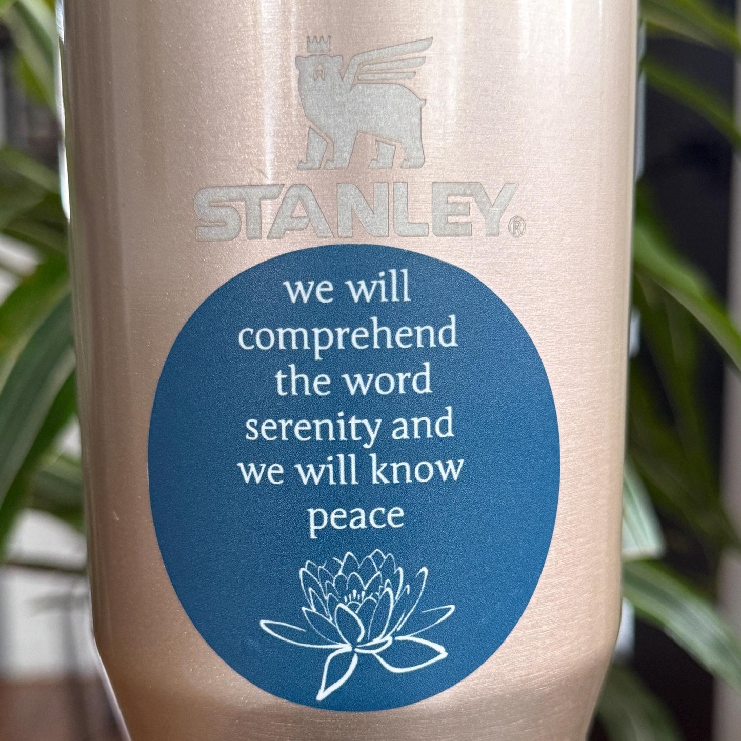 Stanley tumbler with a 2.5" blue sticker with a quote from the big book of AA, "we will comprehend the word serenity and we will know peace" and a lotus flower.