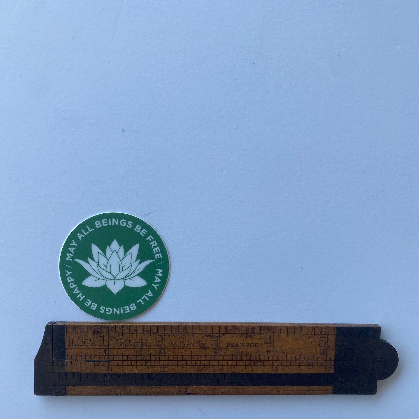 Green circular sticker with white lotus flower design and text on a light blue background, next to a wooden ruler.