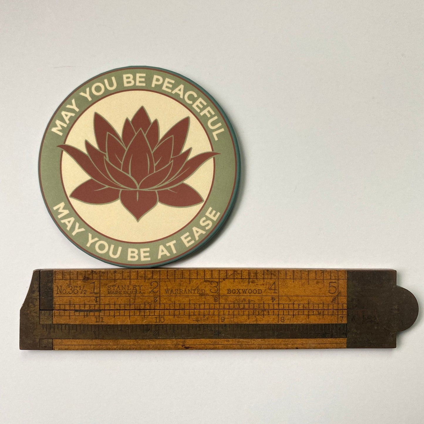 3.5" lotus flower magnet with the metta prayer, 'may you be peaceful may you be at ease' placed above a wooden ruler on a light gray background.