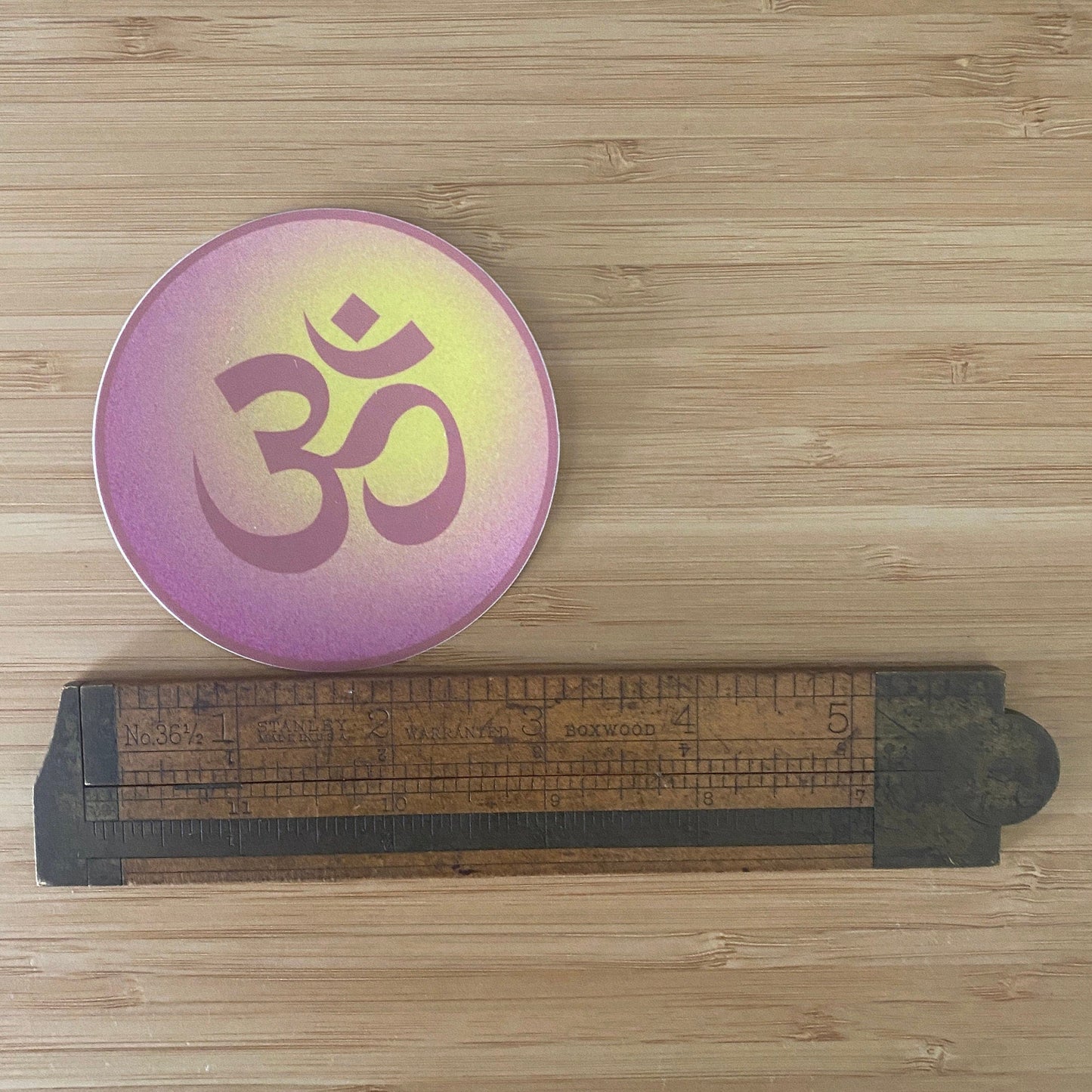 Round pink and gold Om symbol on a wooden surface with a ruler for scale.