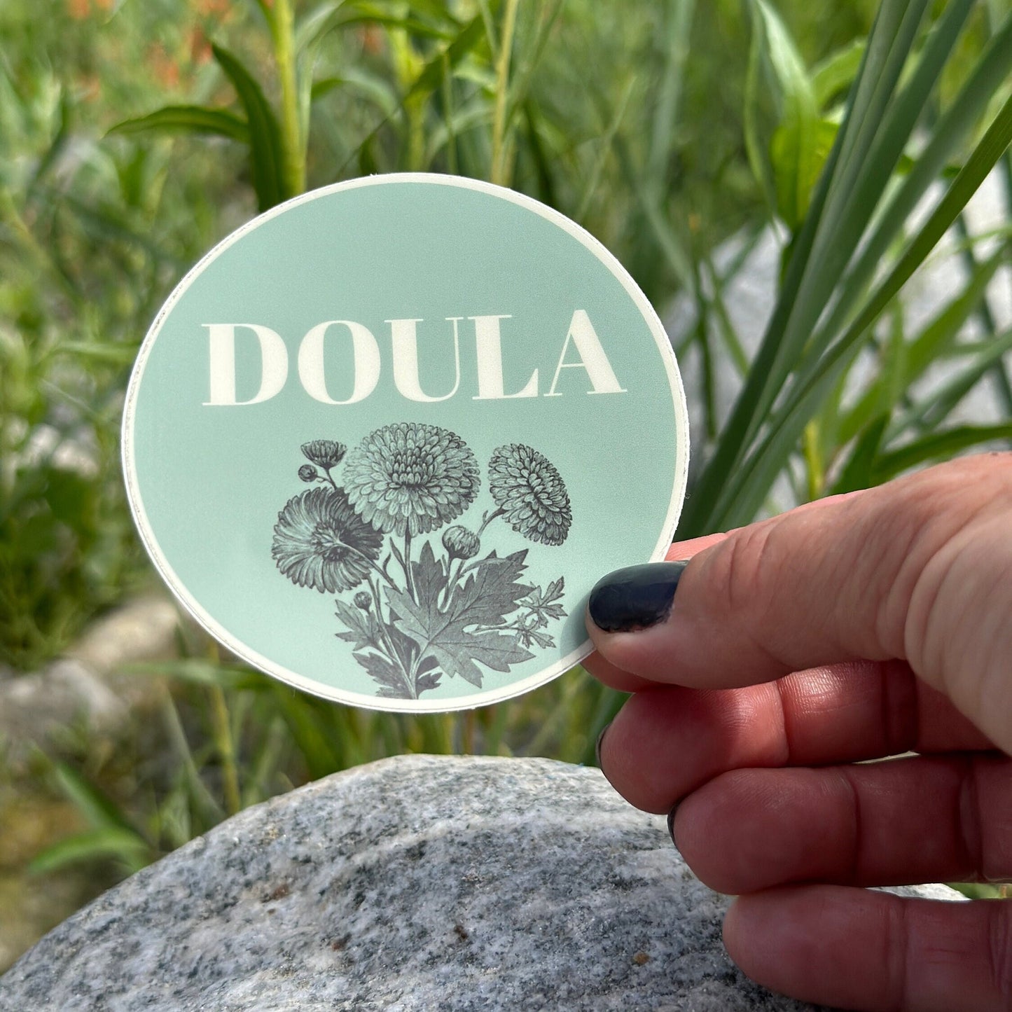 Round green sticker with 'DOULA' text held by a hand against a natural background.